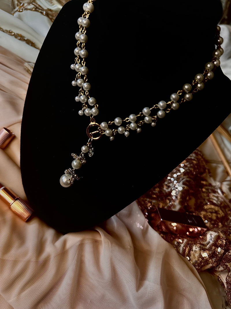 A pearl and gold necklace displayed on a black jewelry bust, surrounded by draped champagne-colored fabric and gold-toned accessories.